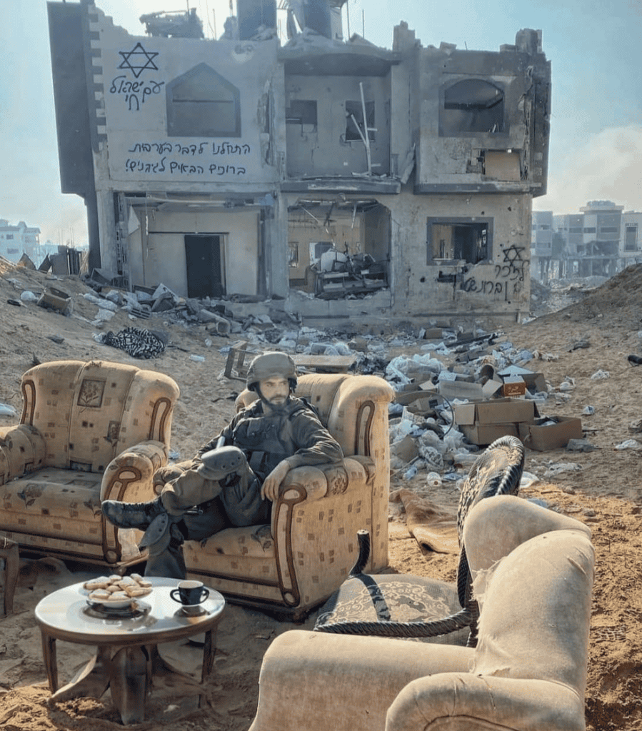 An IDF soldier sitting outside on a living room sofa and coffee table set in front of a bombed-out building. The image has been photoshopped to add cookies to the coffee table and anti-Arabic graffiti to the building.